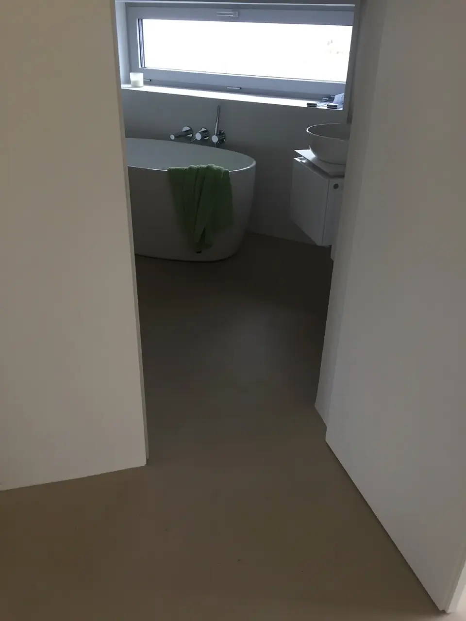 Pohled dveřmi na volně stojící vanu a béžovou stěrkovou podlahu