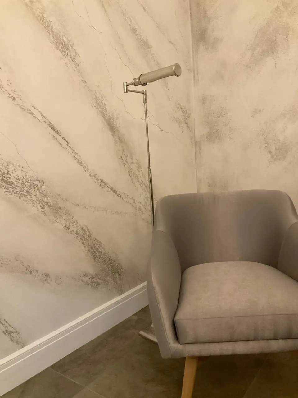 Detail mramorové stěrky s nástěnnou lampou a šedým křeslem